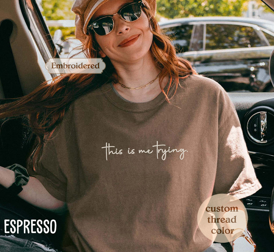 A woman wearing a brown t-shirt with the text "This is me trying" embroidered on it, smiling and looking at the camera.