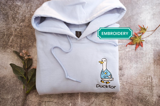 A white hoodie with a cartoon duck printed on it, along with some leaves and flowers, is displayed on a textured surface.