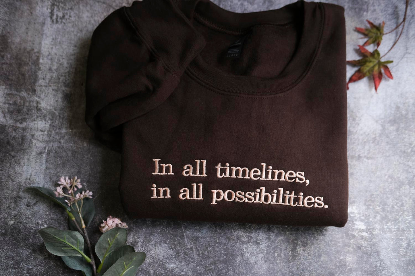 a dark brown t-shirt with the text "In all timelines, in all possibilities" embroidered on it, along with a small plant and some leaves.
