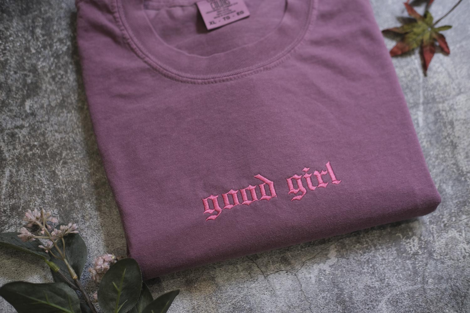 A purple t-shirt with the text "good girl" printed in pink embroidery, along with some leaves and flowers on a textured surface.