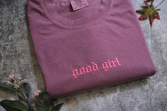 A purple t-shirt with the text "good girl" printed in pink embroidery, along with some leaves and flowers on a textured surface.