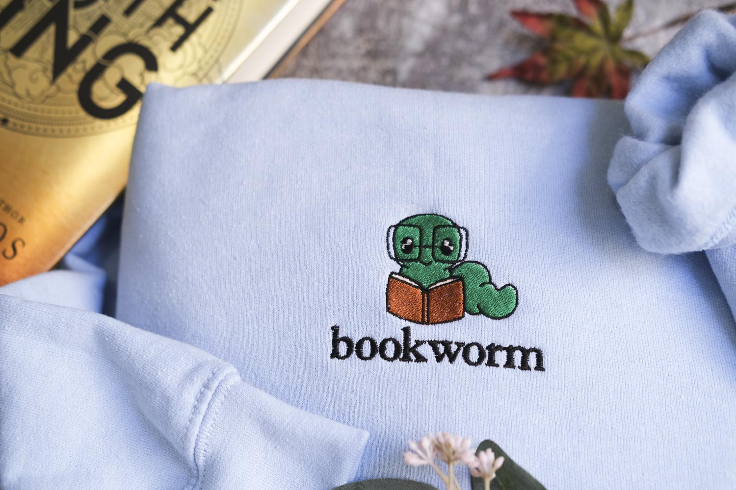 a light blue sweatshirt with a small green book worm wearing glasses and holding a book, along with the word "bookworm" printed on it.