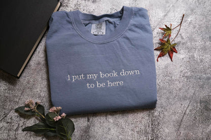 a blue t-shirt with the text "I put my book down to be here" printed on it, along with some leaves and a book.