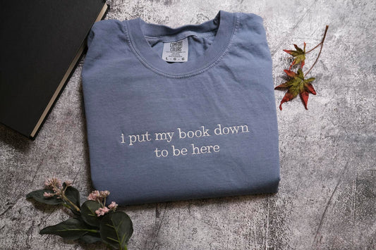 a blue t-shirt with the text "I put my book down to be here" printed on it, along with some leaves and a book.