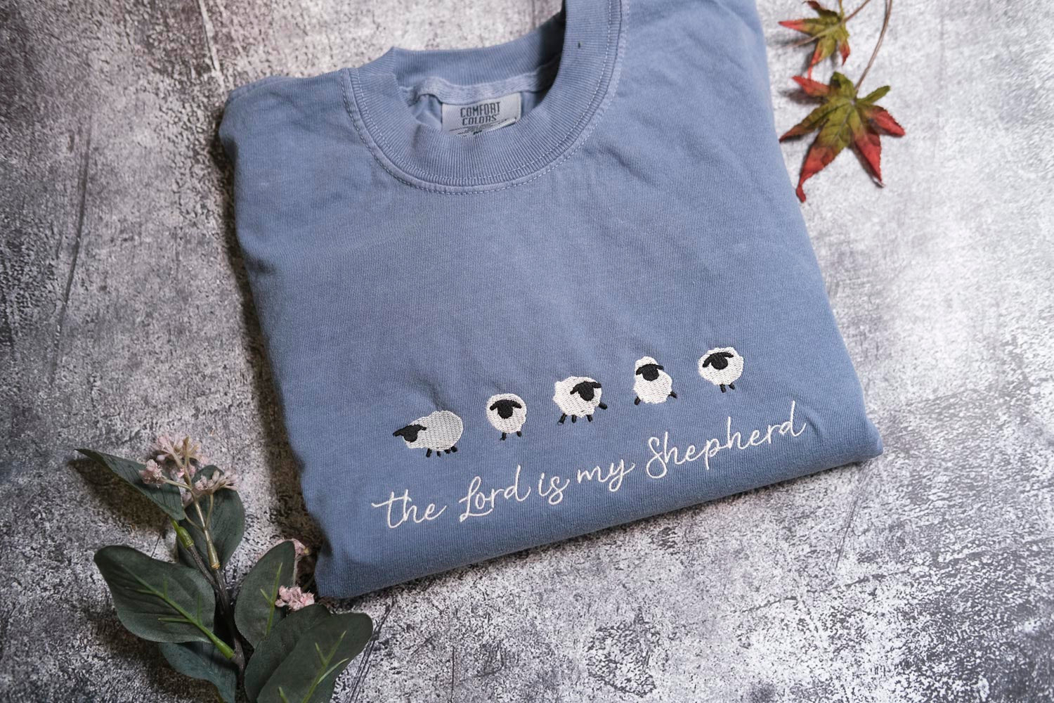 A blue t-shirt with a white sheep embroidered design and the text "The Lord is my Shepherd" printed on it, along with a small leaf and a flower.