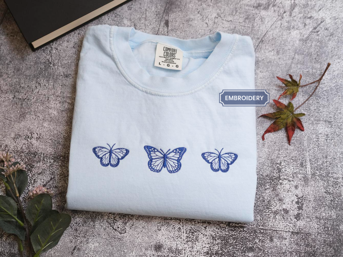 a white t-shirt with three blue butterflies printed on it, placed on a gray surface alongside some leaves and a book.