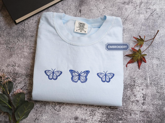 a white t-shirt with three blue butterflies printed on it, placed on a gray surface alongside some leaves and a book.