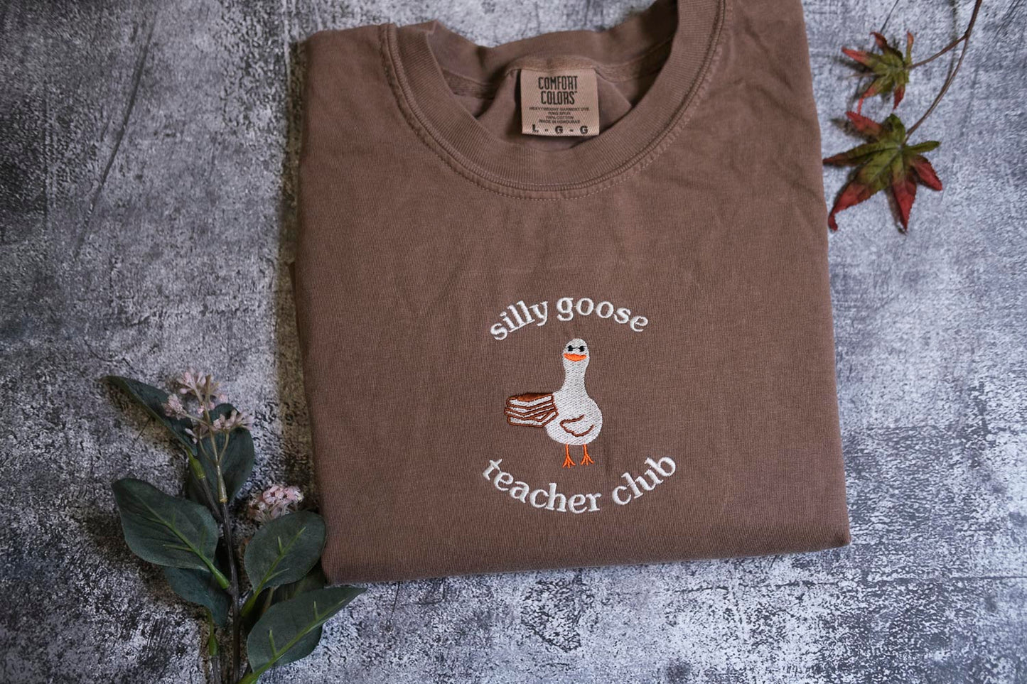 a brown t-shirt with a white goose and the text "silly goose teacher club" printed on it, along with a small label.