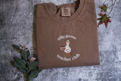 a brown t-shirt with a white goose and the text "silly goose teacher club" printed on it, along with a small label.