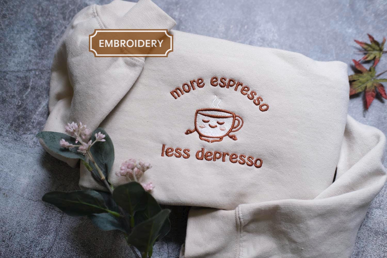 a white sweater with an embroidered design on it, which says "more espresso less depression". The sweater is placed on a gray surface, and there are some flowers and leaves nearby.
