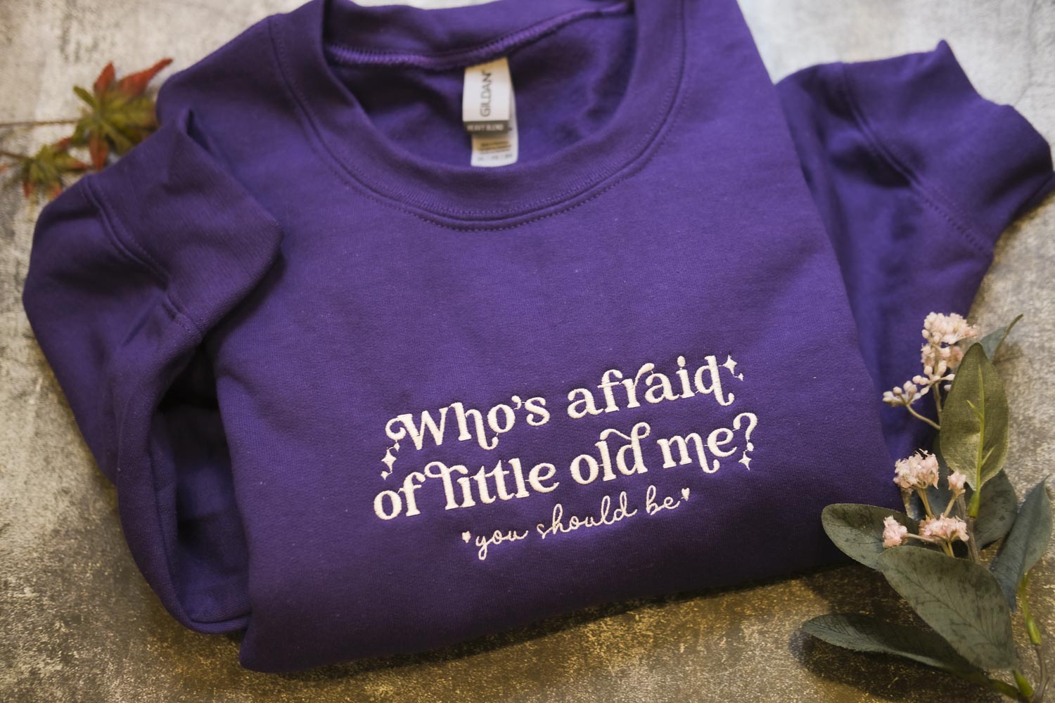 A purple sweatshirt with the text "Who's afraid of little old me?" printed on it, along with a small plant and some flowers.