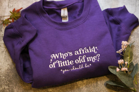 A purple sweatshirt with the text "Who's afraid of little old me?" printed on it, along with a small plant and some flowers.