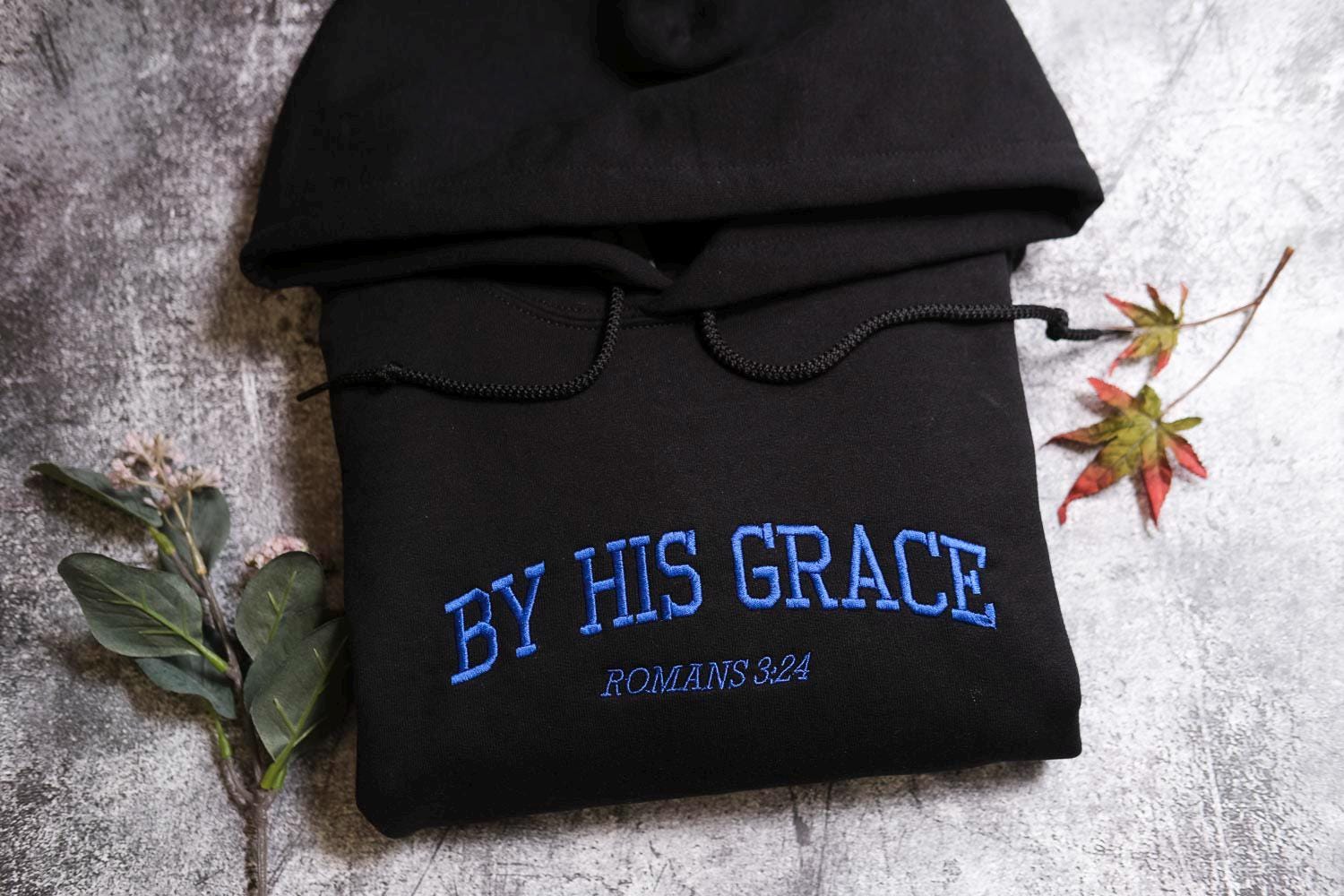 a black hoodie with the text &quot;BY HIS GRACE&quot; and &quot;ROMANS 824&quot; embroidered on it, along with a leaf and a branch.