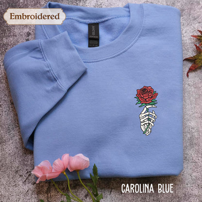 a blue long-sleeved shirt with a rose embroidered on the front, along with a pair of pink flowers. The shirt is laid out on a gray surface, and there is a label visible on the collar.