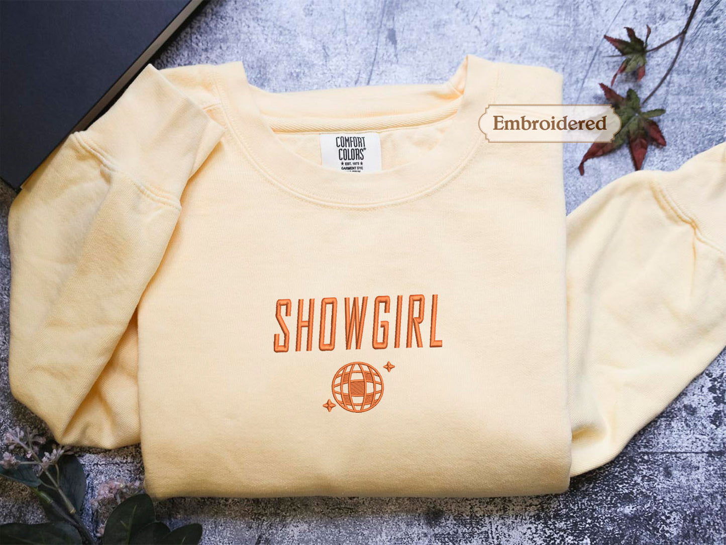 a yellow t-shirt with the text "SHOWGIRL" printed on it, along with a globe design. The t-shirt is folded and placed on a surface, with a label indicating that it is embroidered.