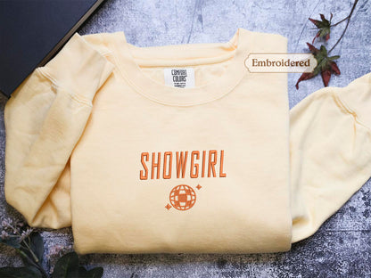 a yellow t-shirt with the text "SHOWGIRL" printed on it, along with a globe design. The t-shirt is folded and placed on a surface, with a label indicating that it is embroidered.