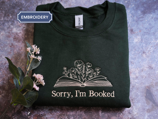 a dark green t-shirt with an embroidered design on it, which says "Sorry, I'm Booked". The t-shirt is placed on a surface with some flowers nearby.