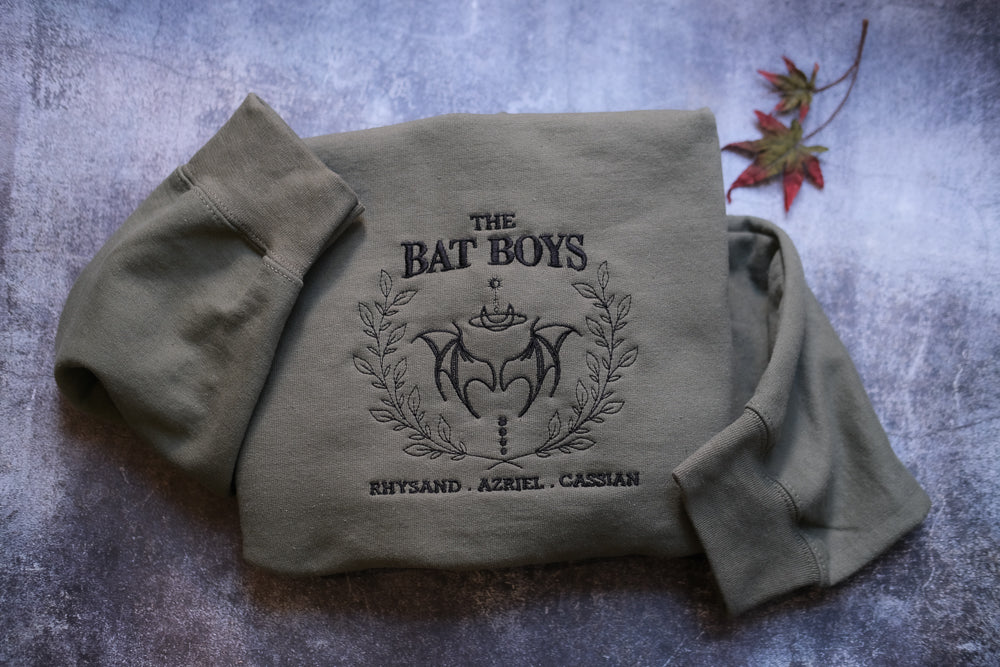 A grey sweatshirt with the text "THE BAT BOYS" and "RHYTHSD. AZIEL. CASSIAN" printed on it, along with a bat and laurel wreath design.