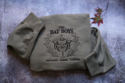 A grey sweatshirt with the text "THE BAT BOYS" and "RHYTHSD. AZIEL. CASSIAN" printed on it, along with a bat and laurel wreath design.