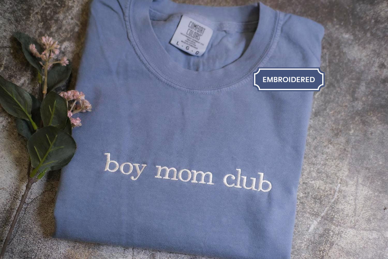 A blue t-shirt with the text "boy mom club" printed on it, along with a label indicating that it is an embroidered item.