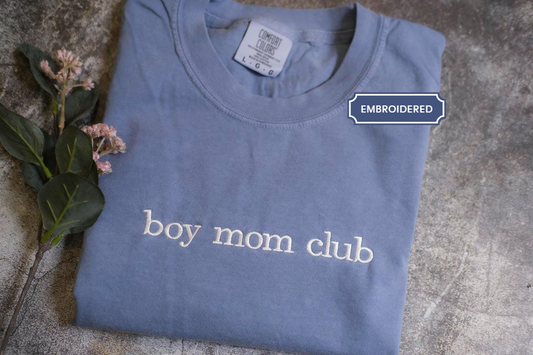 A blue t-shirt with the text "boy mom club" printed on it, along with a label indicating that it is an embroidered item.