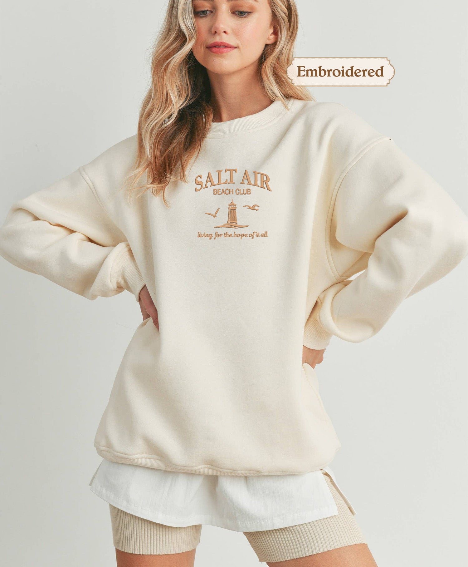 A woman wearing a cream-colored sweater with the text "Salt Air Beach Club" printed on it, standing with her hands on her hips.