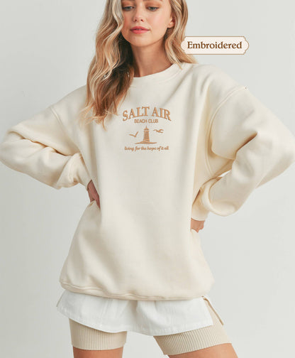 A woman wearing a cream-colored sweater with the text "Salt Air Beach Club" printed on it, standing with her hands on her hips.