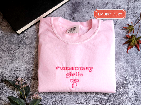 a pink t-shirt with the text "romantic girlie" printed on it, along with a bow design. The t-shirt is folded and placed on a gray surface, with a black book and some leaves in the background.