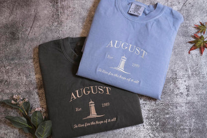 two t-shirts with the word "AUGUST" printed on them, laid out on a textured surface.