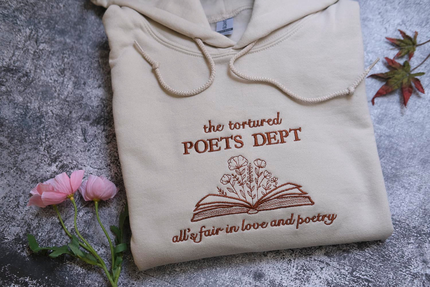 a beige hoodie with a brown embroidered design, which says "the tortured POET'S DEPT all fair in love and poetry". The hoodie is placed on a gray surface, with a small bouquet of pink flowers and a fallen leaf nearby.