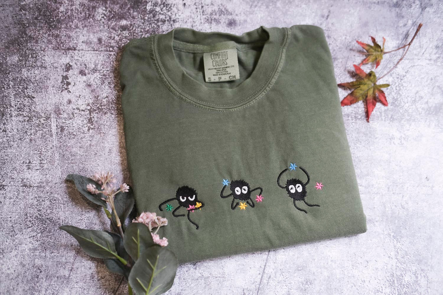 a green t-shirt with three small black figures and flowers embroidered on it, along with some leaves and flowers on a textured surface.