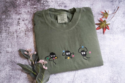 a green t-shirt with three small black figures and flowers embroidered on it, along with some leaves and flowers on a textured surface.