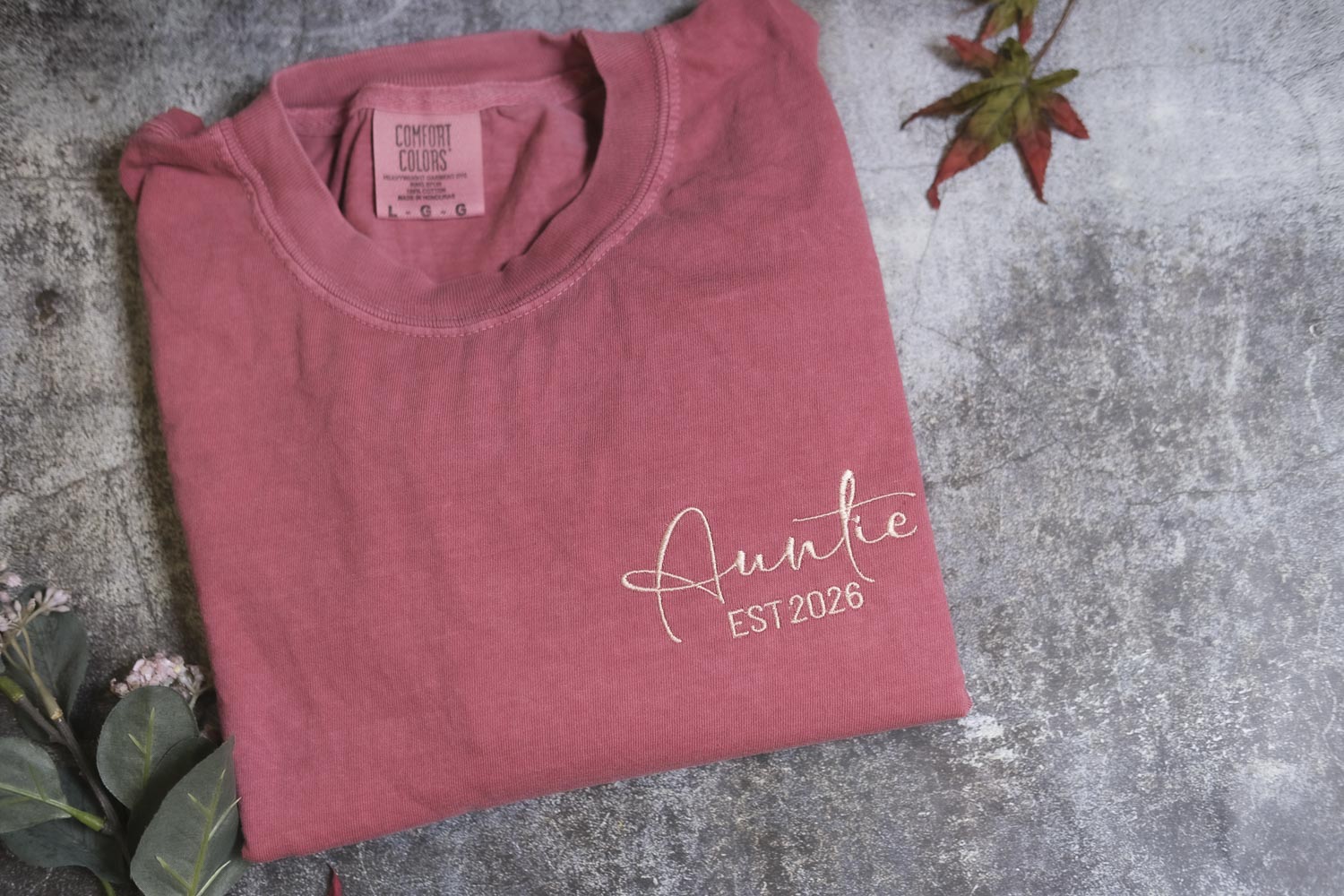 A pink t-shirt with the text "Auntie EST2026" printed on it, along with a small label on the collar. The t-shirt is placed on a gray surface, with a small plant and a red leaf visible in the background.