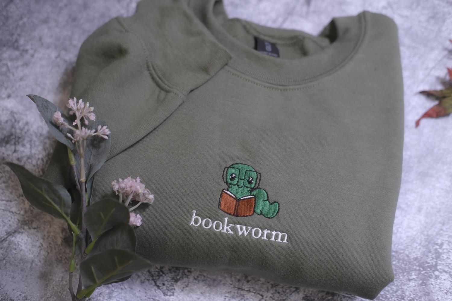 A green sweatshirt with a cartoon image of a bookworm and the word "bookworm" printed on it, along with some pink flowers and a leaf.