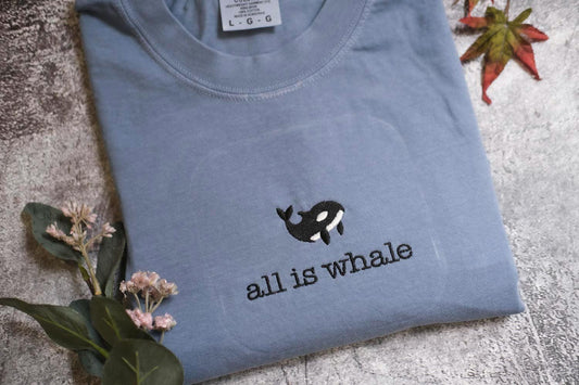A blue t-shirt with a black and white whale embroidered on it, along with some flowers and a leaf, all placed on a gray surface.