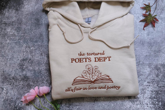 a beige hoodie with a brown embroidered design and text, along with some pink flowers and a leaf, all placed on a textured gray surface.