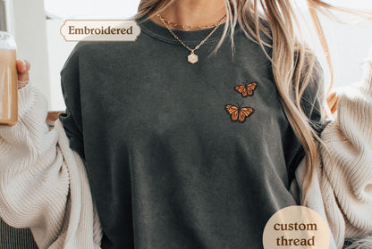 A person wearing a gray t-shirt with two embroidered butterflies on the front, along with a necklace and a sweater.