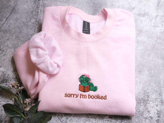 A pink sweatshirt with a cartoon image of a green caterpillar and the text "Sorry I'm bookeded" printed on it.