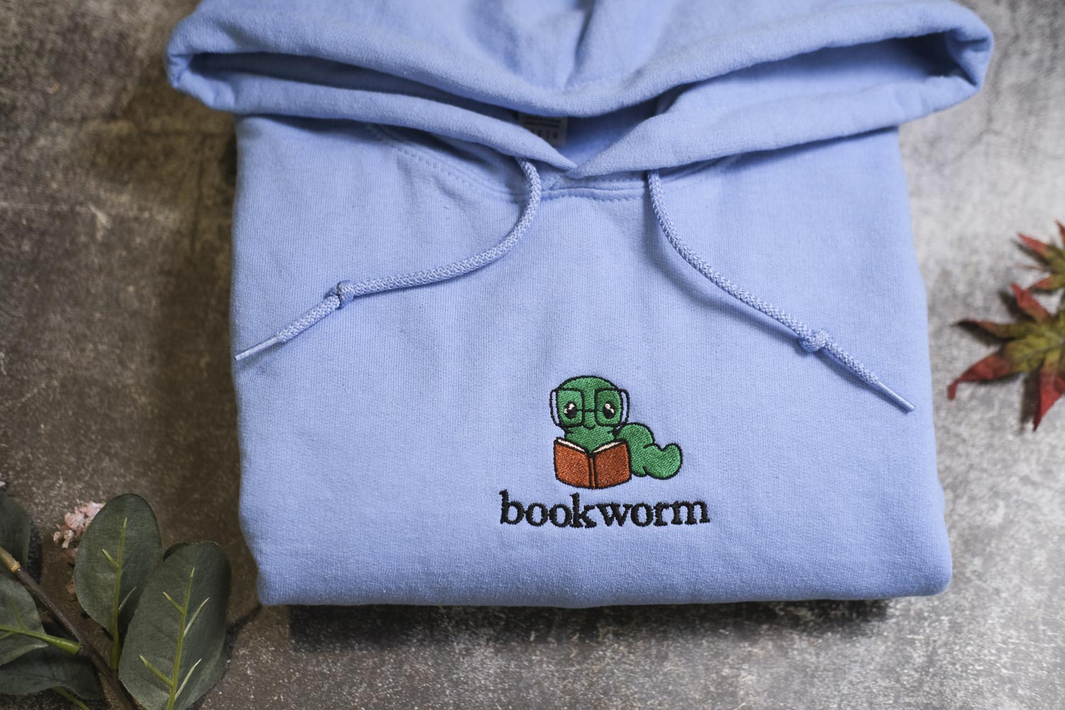 A light blue hoodie with a cartoon image of a book worm and the text "bookworm" printed on it, along with some leaves and a small plant.