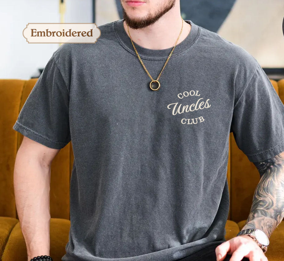 A man wearing a gray t-shirt with the text "Cool Uncle Club" embroidered on it, sitting on a couch.