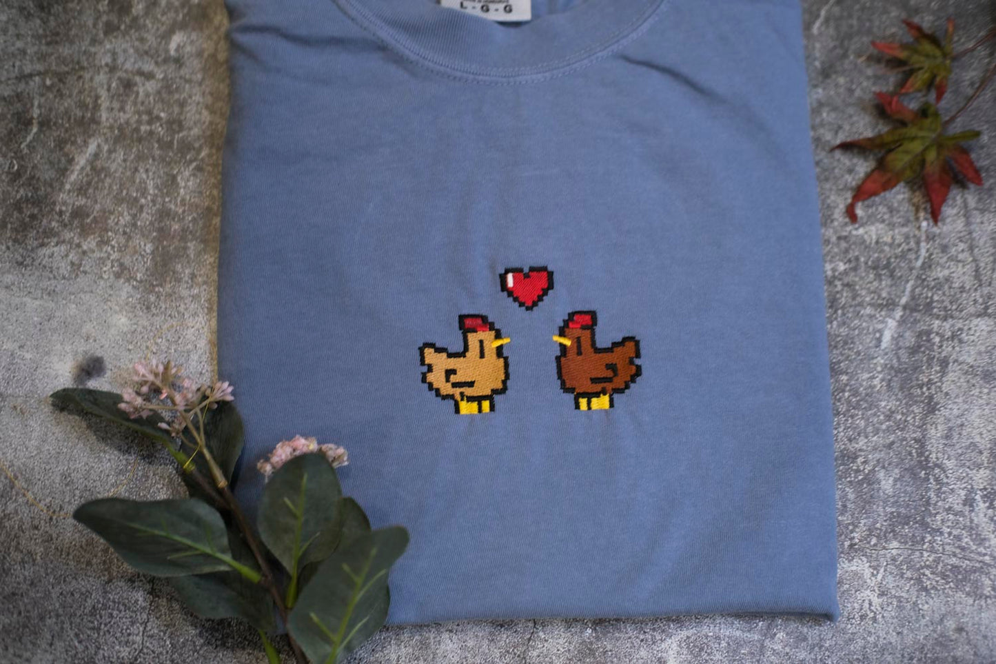 a blue t-shirt with a pixelated design of two cartoon chickens, one brown and one yellow, with a heart above them. The t-shirt is placed on a gray surface, and there are some flowers and leaves in the background.