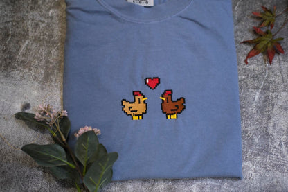 a blue t-shirt with a pixelated design of two cartoon chickens, one brown and one yellow, with a heart above them. The t-shirt is placed on a gray surface, and there are some flowers and leaves in the background.