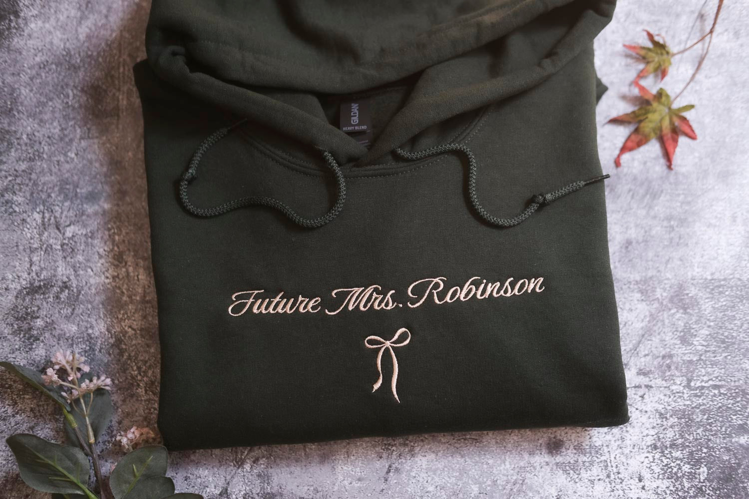 A dark green hoodie with the text "Future Mrs. Robinson" printed on it, along with a bow symbol, is displayed on a gray surface.
