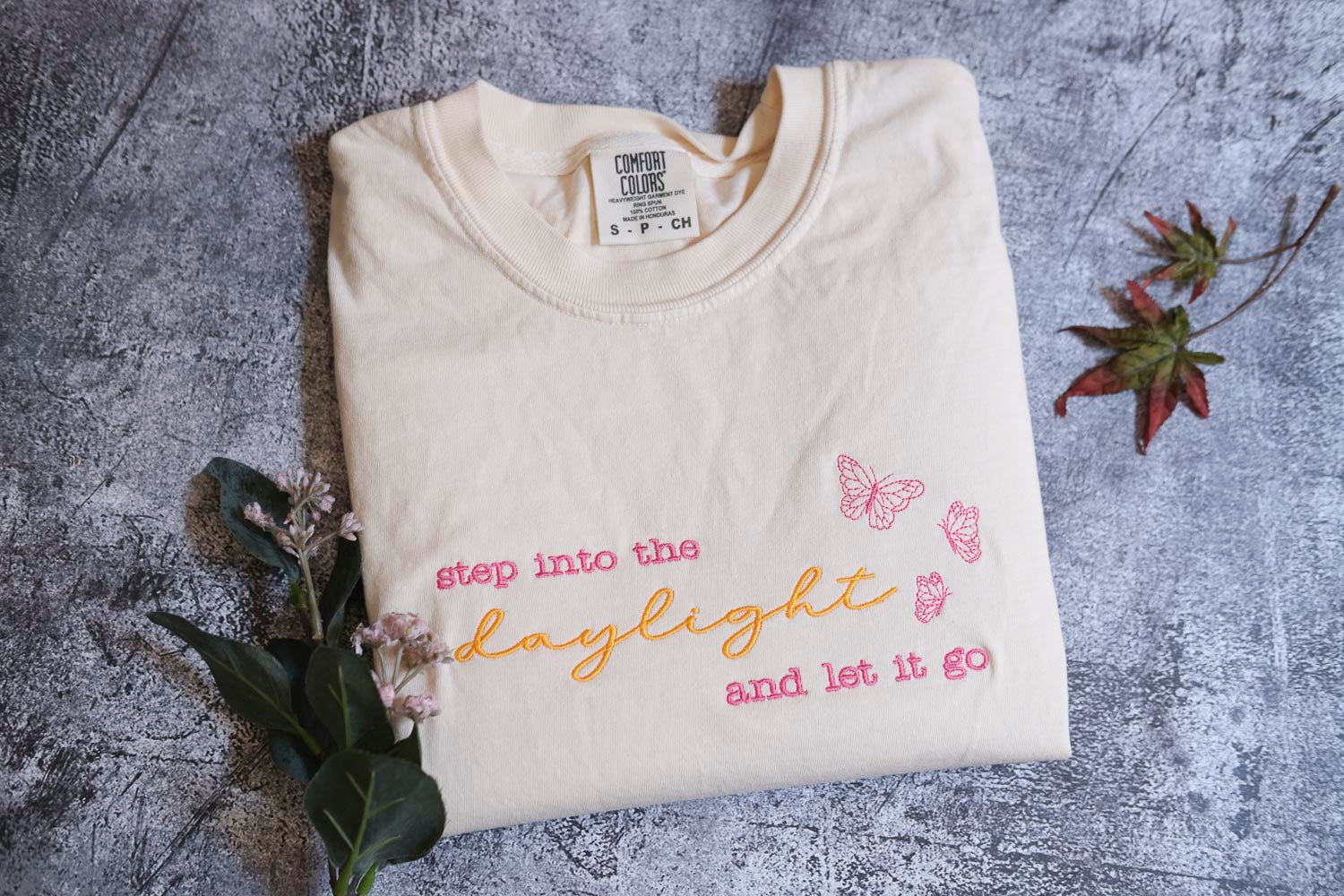 A white t-shirt with text and butterflies printed on it, along with some flowers and leaves, is displayed on a gray surface.