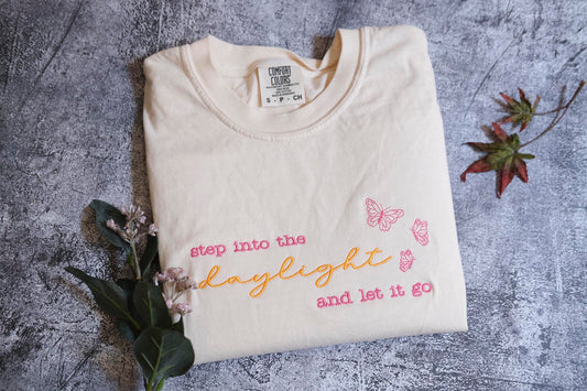 A white t-shirt with text and butterflies printed on it, along with some flowers and leaves, is displayed on a gray surface.