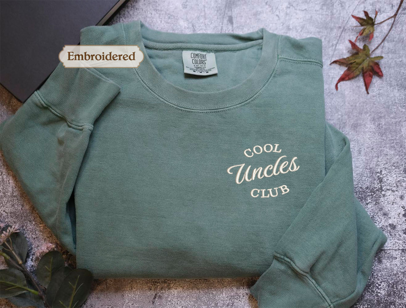 A green long-sleeved shirt with the embroidered text "Cool Uncle Club" on the front, placed on a gray surface with a leaf and a small object in the background.