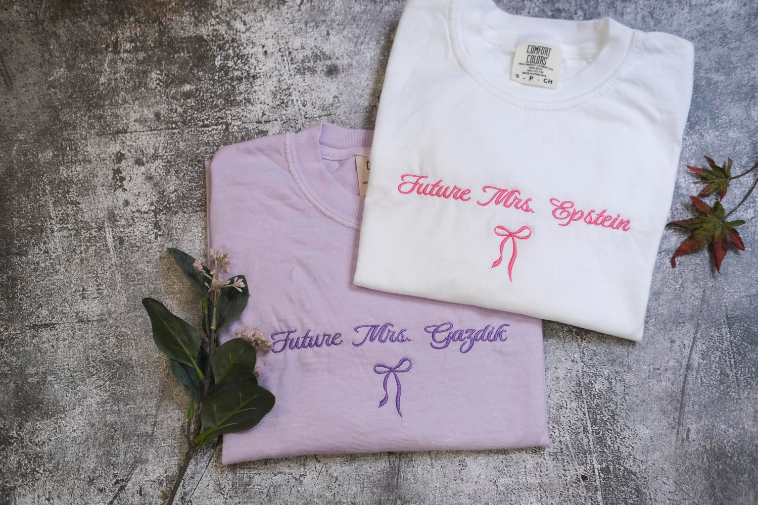 two white t-shirts with the text "Future Mrs. Einstein" printed on them, along with a bow. The t-shirts are placed on a gray surface, and there are some flowers and leaves nearby.
