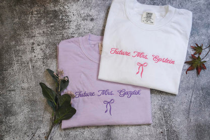 two white t-shirts with the text "Future Mrs. Einstein" printed on them, along with a bow. The t-shirts are placed on a gray surface, and there are some flowers and leaves nearby.
