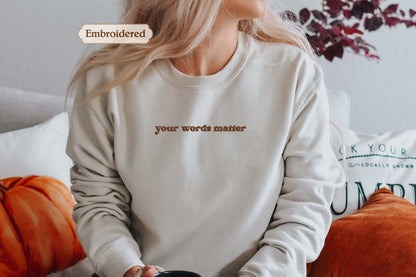 A woman wearing a white sweatshirt with the text "your words matter" embroidered on it, sitting on a couch with orange pillows.