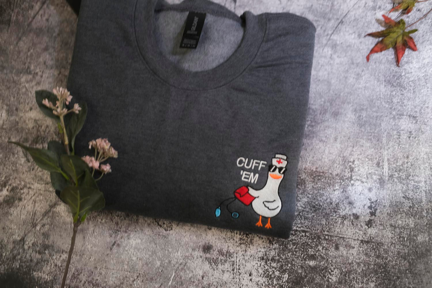a gray t-shirt with a cartoon duck and the text "CUFF 'EM" printed on it, placed on a gray surface alongside some flowers.